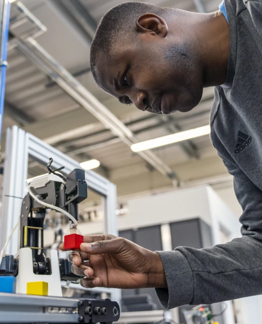 Student adjusting a robotic arm to pick up a red block, focusing intently on precision in a mechatronics workshop. Student adjusting a robotic arm to pick up a red block, focusing intently on precision in a mechatronics workshop.